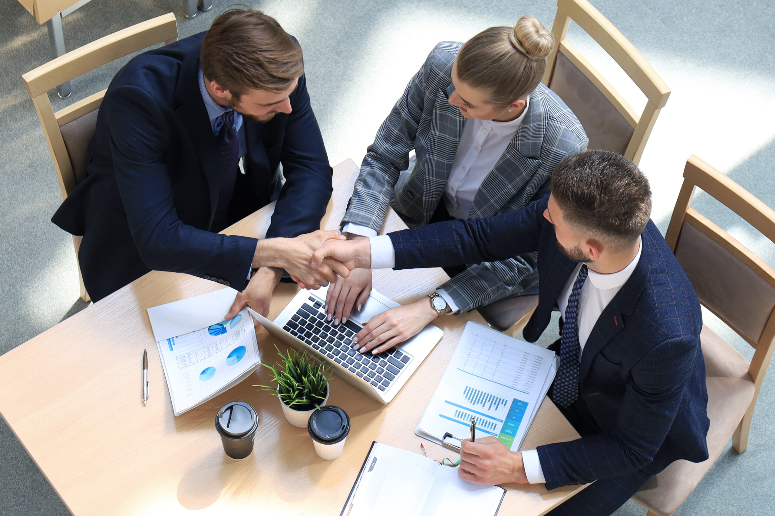 Top view of two men sitting at the desk and shaking hands while young woman looking at them and smiling